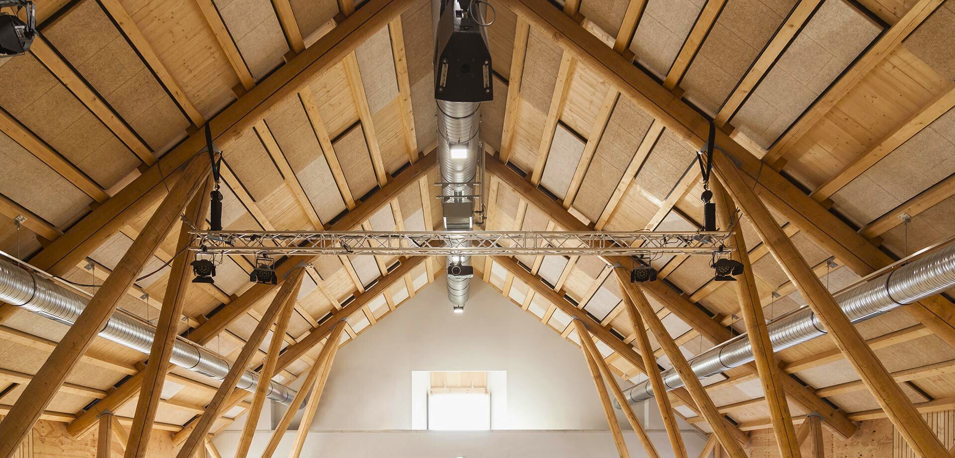 Ein Holzdach mit massiven, sichtbaren Holzträgern und Lüftungsrohren, die von der Decke hängen. Beleuchtungs- und Audiogeräte sind an einem Metallgerüst montiert.