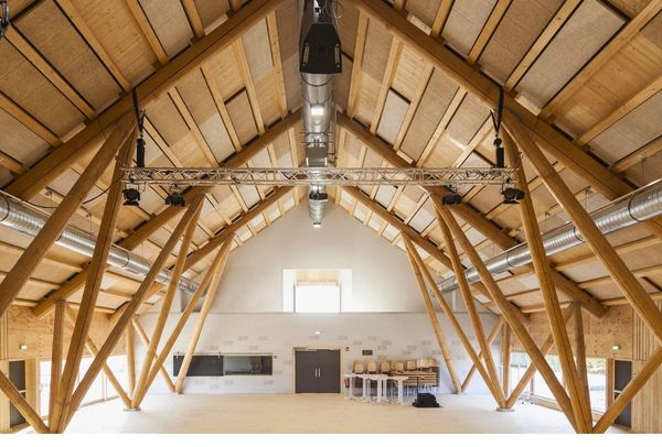 Ein Innenraum mit einem hohen Dachboden, der von sichtbaren, massiven Holzstreben gestützt wird. Die Decke und Streben sind aus hellem Holz, was dem Raum ein rustikales, aber einladendes Ambiente verleiht. Ein Kronleuchter hängt in der Mitte und beleuchtet den Raum.