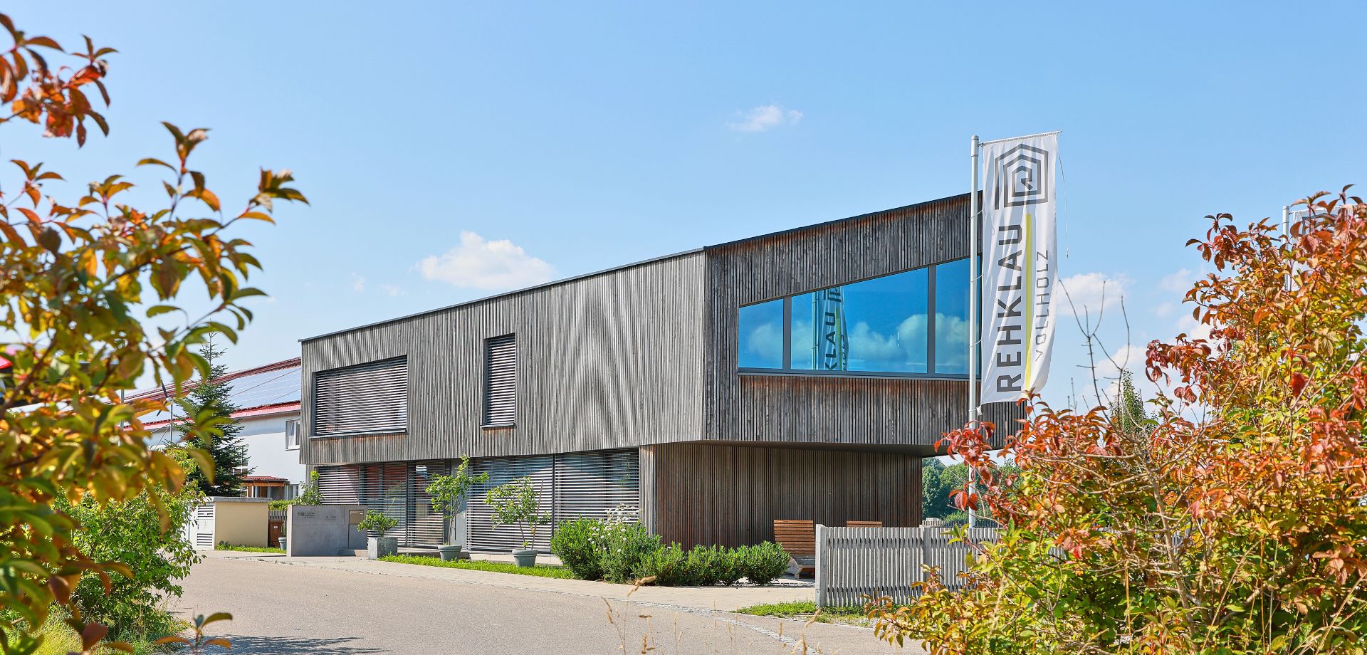 Modernes Bürogebäude aus Rehklau Vollholz mit Rehklau-Fahne Ein modernes, zweistöckiges Gebäude mit einer Fassade aus Holz, das den Namen "Rehklau" auf einer Fahne zeigt. Die großen Fenster reflektieren den blauen Himmel. Umgeben von Büschen und grüner Vegetation, steht das Gebäude in einer ruhigen Umgebung.